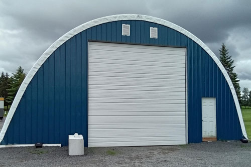 A blue, semi-circular barn with a large white garage door and a smaller white entry door on a gravel-covered ground, under a cloudy sky